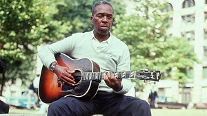 Black and white photograph of Skip James seated on a park bench playing an acoustic guitar.