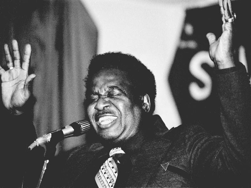 Black and white close-up photograph of Roy Brown singing into a microphone with both hands raised.
