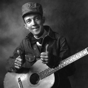 Studio portrait of Jimmie Rodgers smiling with a guitar and wearing a railroad cap.