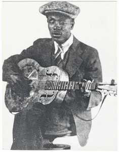 Studio photograph of Blind Boy Fuller seated and playing a resonator guitar.