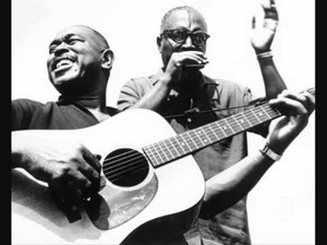 Sonny Terry plays harmonica beside Brownie McGhee playing guitar on an outdoor stage.