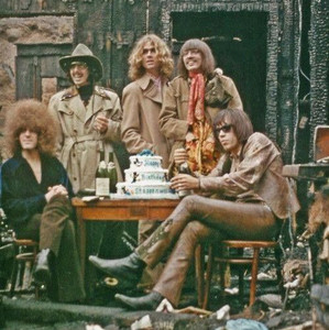 Members of Steppenwolf pose around a birthday cake in a burned-out outdoor setting.