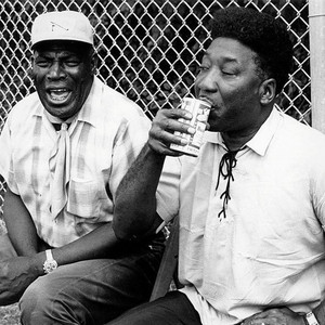 Howlin' Wolf and Muddy Waters seated outdoors beside a chain-link fence.