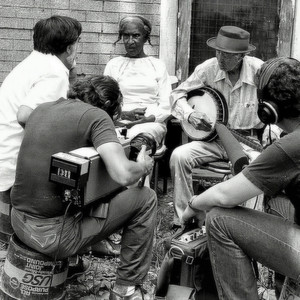 Documentary crew records Dink Roberts playing banjo outdoors beside his seated wife.