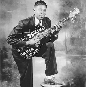 Elderly Gus Cannon seated indoors playing banjo, with a woman seated in the background.