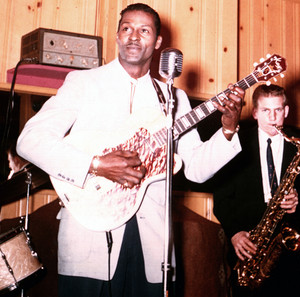 Chuck Berry performing with guitar in a studio setting, with a saxophonist behind him.