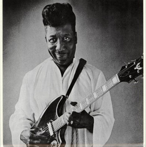 Muddy Waters smiling in a studio while holding a Guild Thunderbird guitar.
