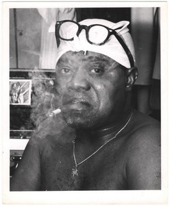 Louis Armstrong seated shirtless backstage, smoking with a towel wrapped around his head.