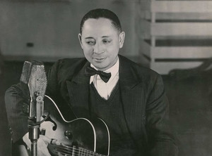 Lonnie Johnson seated in a studio portrait with guitar beside a large microphone.