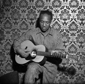 Josh White seated indoors playing acoustic guitar against an ornate wall.