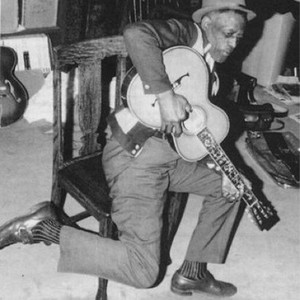 Furry Lewis seated indoors, playing guitar while hooking one leg over a chair back.